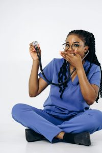 Black female doctor in blue scrubs holding a stethoscope while seated on a white background, smiling playfully.
