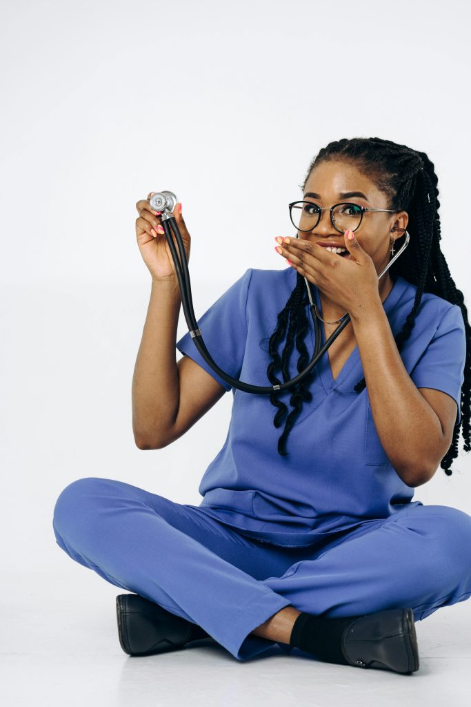Black female doctor in blue scrubs holding a stethoscope while seated on a white background, smiling playfully.